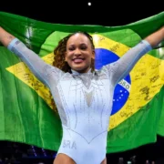 Rebeca Andrade sorri com os braços erguidos, segurando uma bandeira do Brasil atrás do corpo, durante celebração em uma arena de ginástica artística.