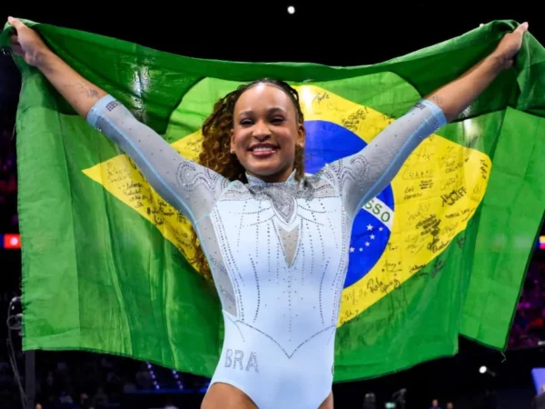 Rebeca Andrade sorri com os braços erguidos, segurando uma bandeira do Brasil atrás do corpo, durante celebração em uma arena de ginástica artística.