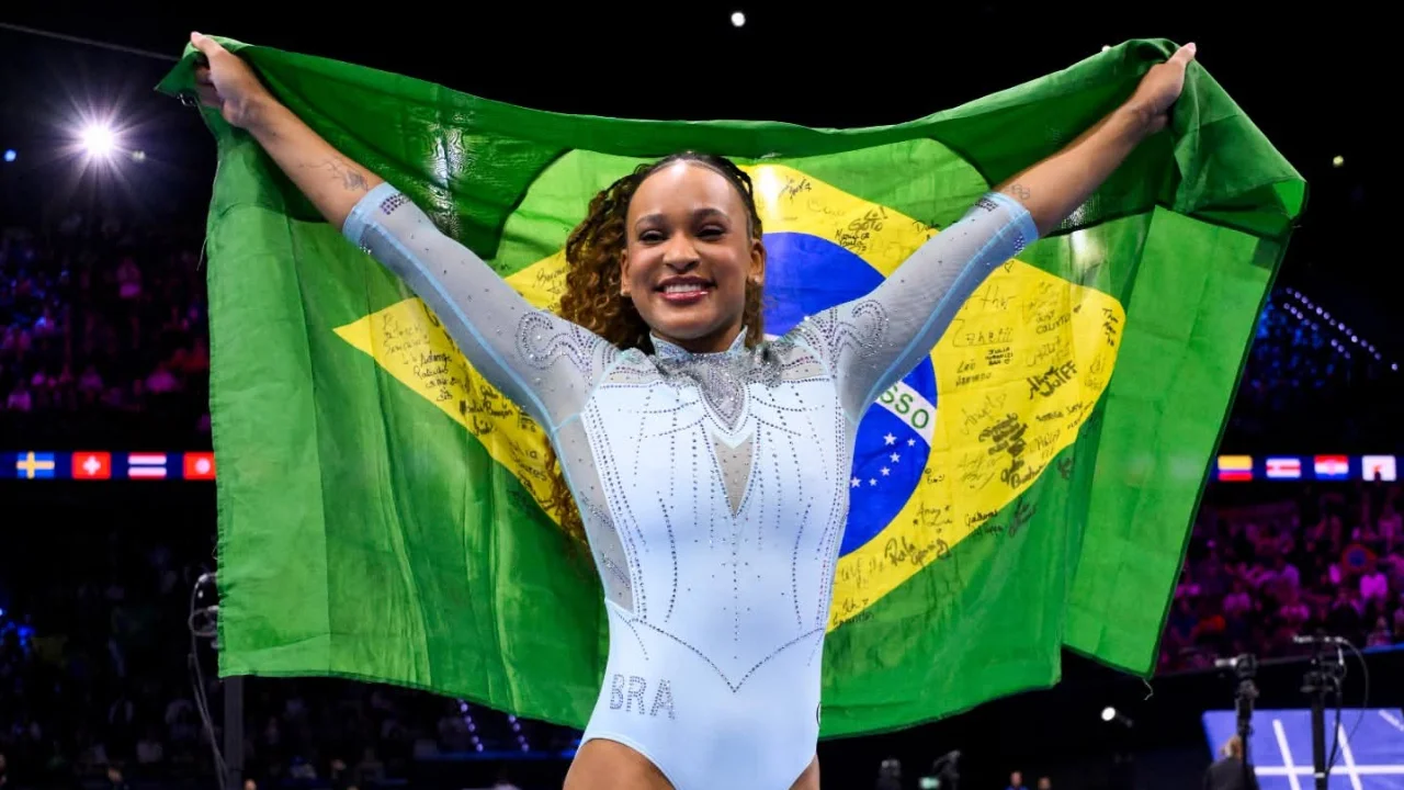 Rebeca Andrade sorri com os braços erguidos, segurando uma bandeira do Brasil atrás do corpo, durante celebração em uma arena de ginástica artística. Rebeca Andrade sorri com os braços erguidos, segurando uma bandeira do Brasil atrás do corpo, durante celebração em uma arena de ginástica artística.