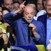 Presidente Lula discursa ao microfone com o braço levantado, cercado por apoiadores e diante de uma bandeira do Brasil, após sua reeleição.