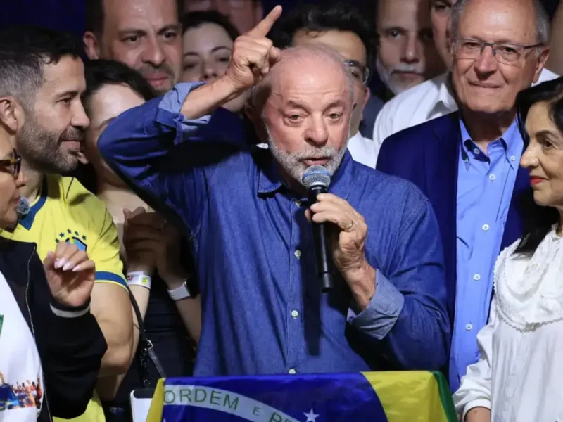 Presidente Lula discursa ao microfone com o braço levantado, cercado por apoiadores e diante de uma bandeira do Brasil, após sua reeleição.