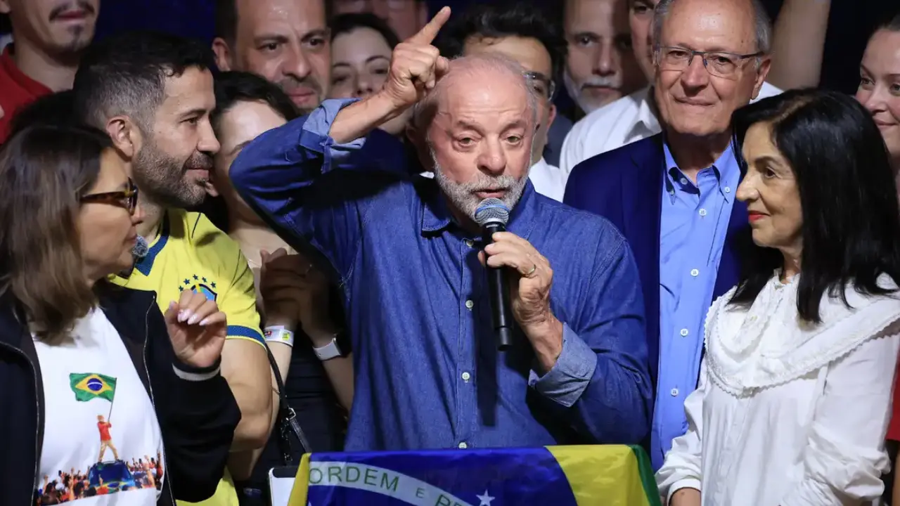 Presidente Lula discursa ao microfone com o braço levantado, cercado por apoiadores e diante de uma bandeira do Brasil, após sua reeleição. Presidente Lula discursa ao microfone com o braço levantado, cercado por apoiadores e diante de uma bandeira do Brasil, após sua reeleição.