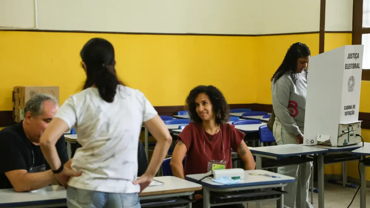 Eleitora utiliza cabine de votação da Justiça Eleitoral enquanto mesários atendem outras pessoas em uma seção eleitoral.