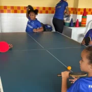Raquete e bola de tênis de mesa sobre uma mesa azul, em ambiente interno.
