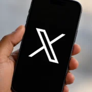 Mão segurando um smartphone com o logotipo branco da rede social X sobre a tela preta, em primeiro plano e com fundo neutro desfocado.