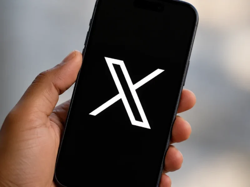 Mão segurando um smartphone com o logotipo branco da rede social X sobre a tela preta, em primeiro plano e com fundo neutro desfocado.