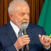 Presidente Lula discursa ao microfone, usando terno azul e gravata, diante de bandeiras do Brasil.