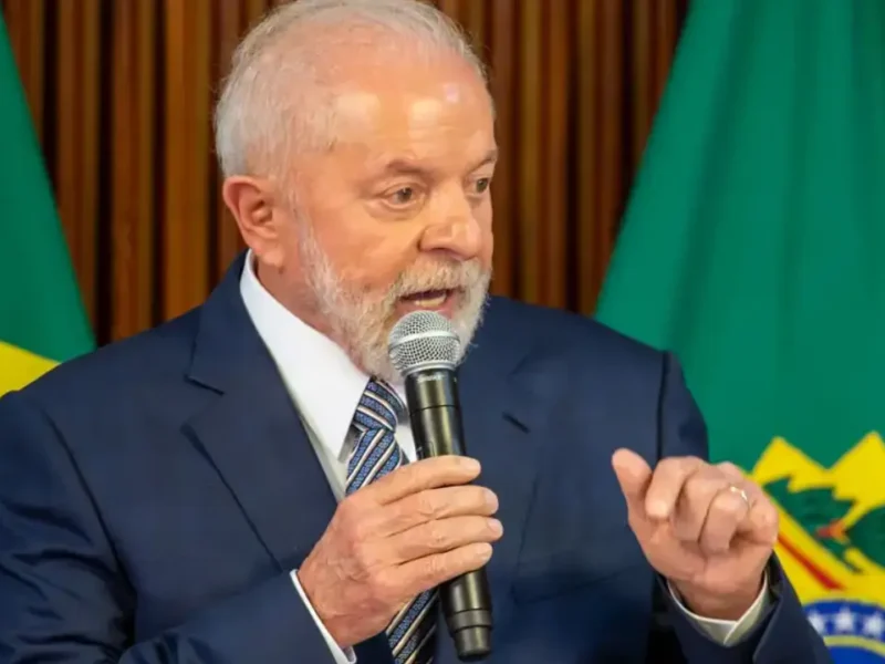 Presidente Lula discursa ao microfone, usando terno azul e gravata, diante de bandeiras do Brasil.