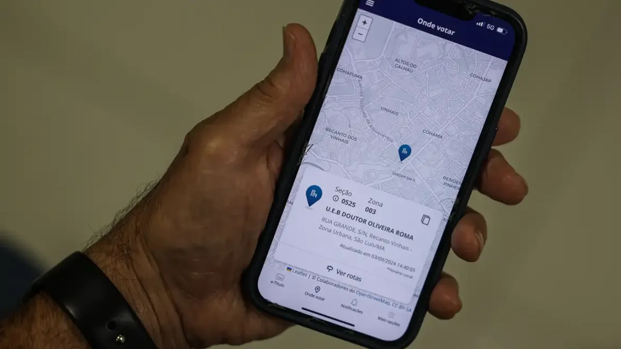 Pessoa segura celular com aplicativo e-Título aberto na função Onde votar, mostrando mapa e endereço da seção eleitoral. Pessoa segura celular com aplicativo e-Título aberto na função Onde votar, mostrando mapa e endereço da seção eleitoral.