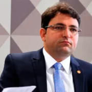 Homem de cabelos escuros e óculos, usando terno azul-marinho, camisa branca e gravata azul-clara, diante de um painel geométrico.