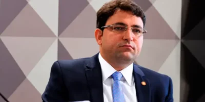 Homem de cabelos escuros e óculos, usando terno azul-marinho, camisa branca e gravata azul-clara, diante de um painel geométrico.